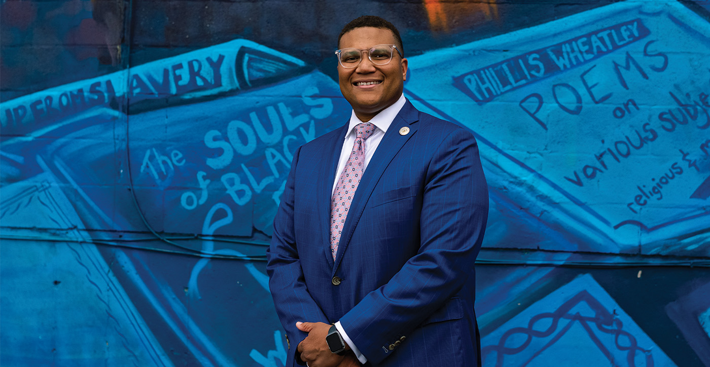 Photo of Brandon Taylor in front of community mural, looking at the camera and smiling